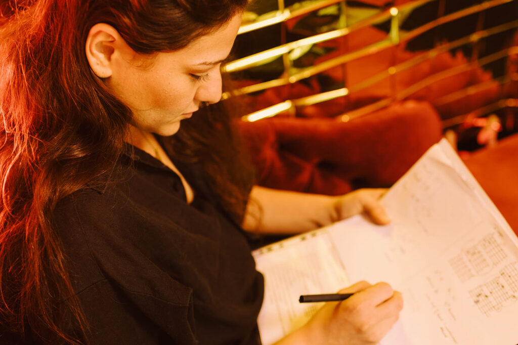 Leyla Mei Parker mit goldenem Hintergrund sitzt auf roter Couch mit Songtexten am Komponieren. 